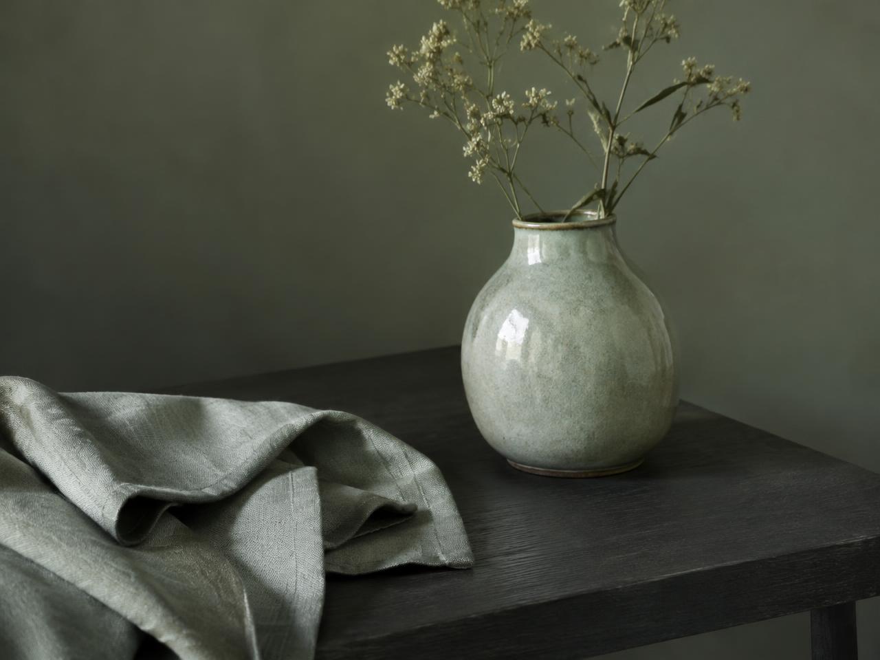 Sage ceramic vase with dried flowers beside folded linen on a dark wooden table